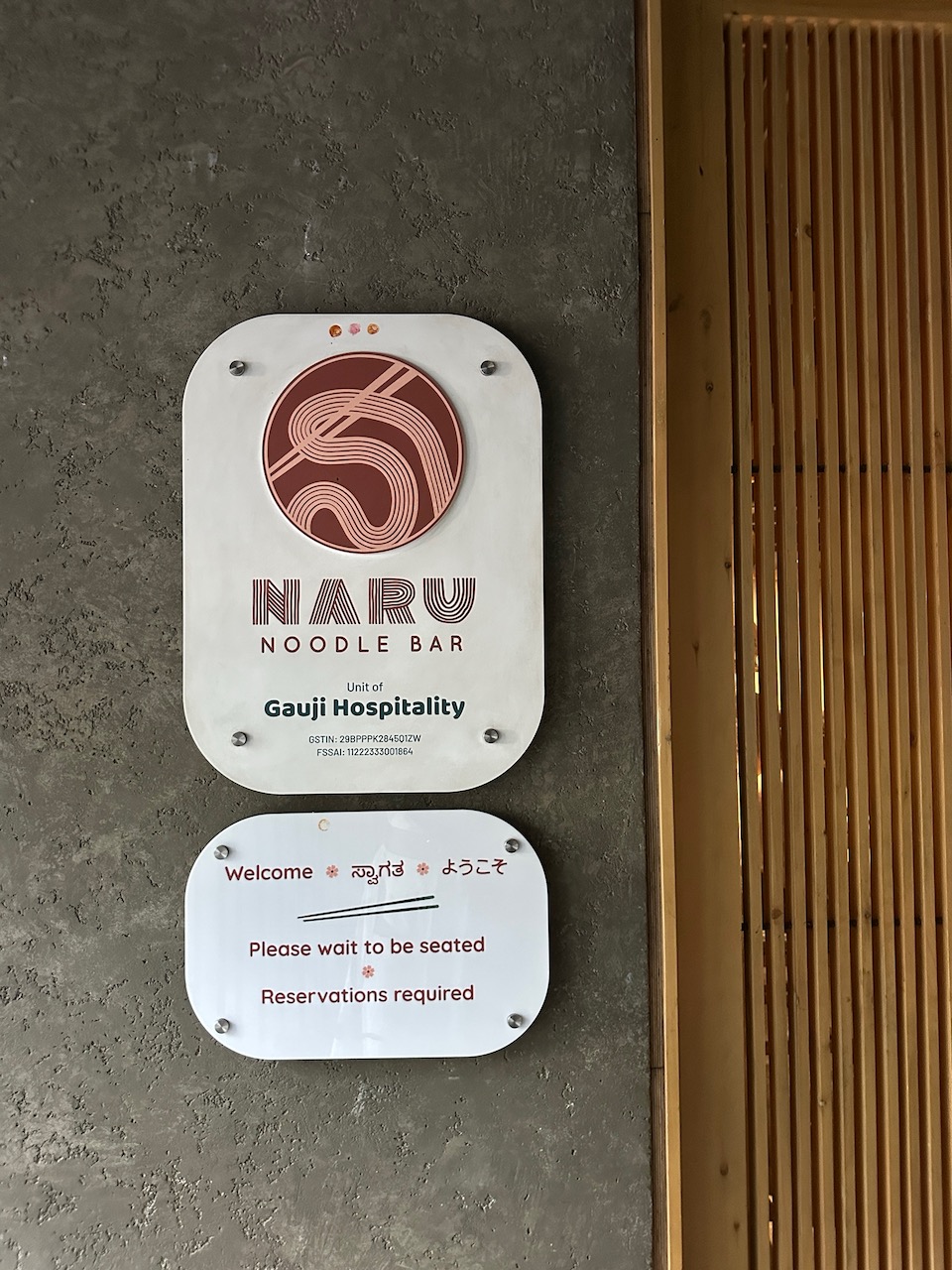 、「5 NOODLE BAR Unitof Gauji Hospitality pitalit GSTIN:2 USTIN:23BPPPK26470ITH 3BPPPK204701ZW PSSAI: 1122223300年04 ခ Welcome 券 ® ಸ್ವಾಗತ # कै ようこそ Please wait Pleasewaittobeseated to be seated # Reservations required ®」というテキストの画像のようです